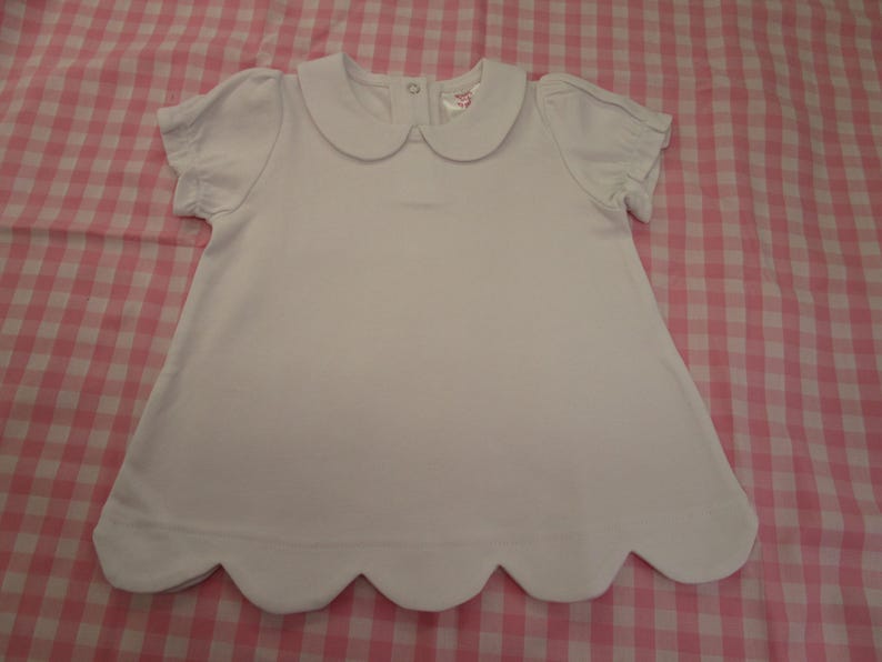 May include: A solid white short-sleeved shirt featuring a rounded Peter Pan collar, gathered sleeve cuffs, and a scalloped hemline. The garment includes a small metal snap closure at the back of the neckline and a visible interior brand label.