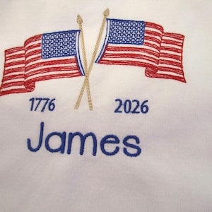May include: White fabric embroidered with two American flags, red and blue, crossed at the poles. The year 1776 is embroidered below one flag, and 2026 below the other. The name "James" is embroidered in blue.