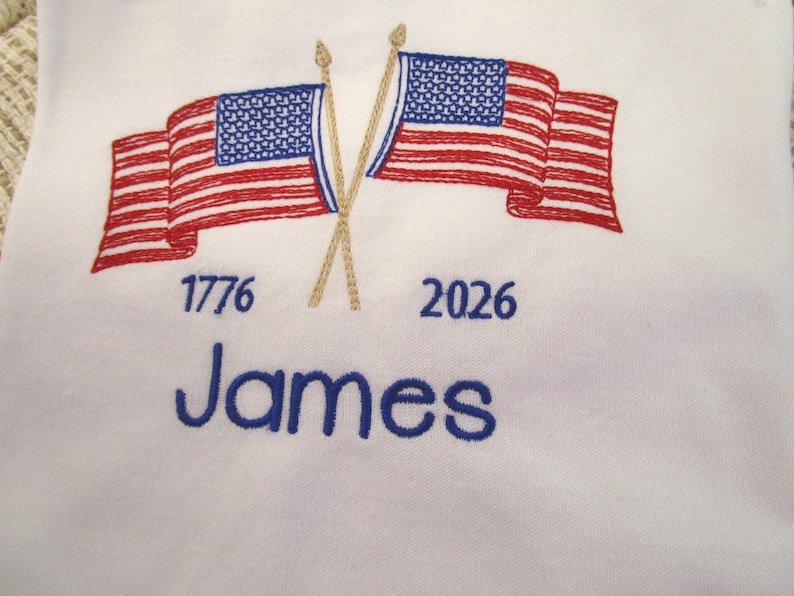 May include: White fabric embroidered with two American flags, red and blue, crossed at the poles. The year 1776 is embroidered below one flag, and 2026 below the other. The name "James" is embroidered in blue.