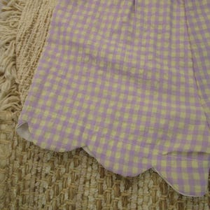 May include: A pair of shorts featuring a light purple and pale yellow gingham check pattern. The hem of the shorts is finished with a distinct scalloped edge detail, providing a curved silhouette along the bottom of the garment fabric.