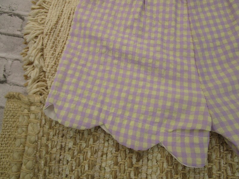 May include: A pair of shorts featuring a light purple and pale yellow gingham check pattern. The hem of the shorts is finished with a distinct scalloped edge detail, providing a curved silhouette along the bottom of the garment fabric.