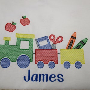 May include: Embroidered design of a green train with a red and yellow car carrying school supplies, including a book, scissors, crayons, and a pencil. The train is decorated with two red apples. The name "James" is embroidered below the train.
