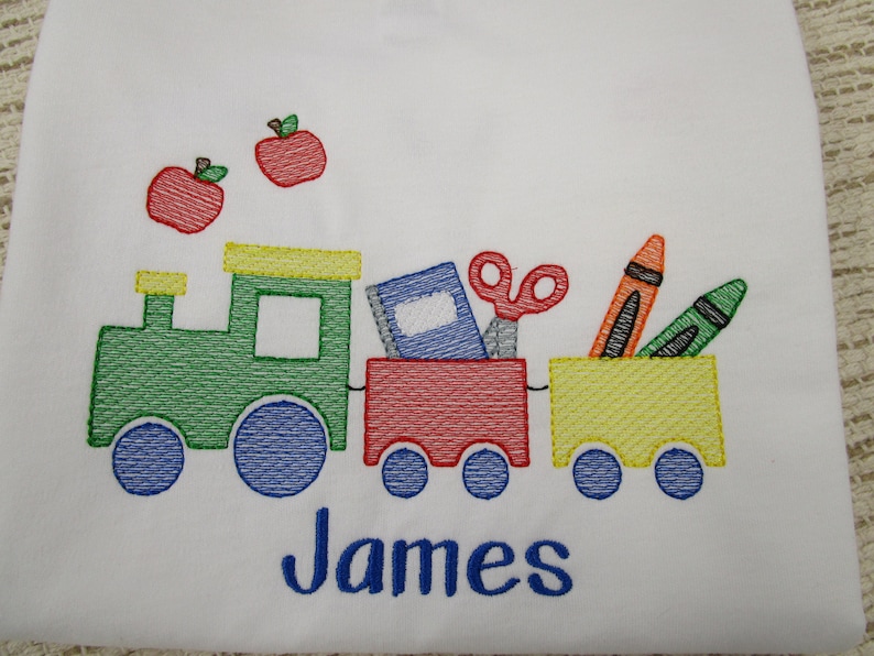 May include: Embroidered design of a green train with a red and yellow car carrying school supplies, including a book, scissors, crayons, and a pencil. The train is decorated with two red apples. The name "James" is embroidered below the train.