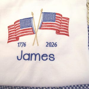 May include: White t-shirt with an embroidered design featuring two American flags crossed at the poles. The flags are red and white striped with blue star fields. The text "1776" and "2026" are above the name "James" in blue.