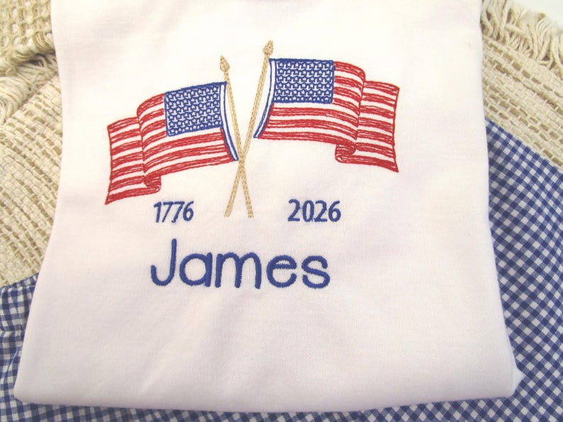 May include: White t-shirt with an embroidered design featuring two American flags crossed at the poles. The flags are red and white striped with blue star fields. The text "1776" and "2026" are above the name "James" in blue.