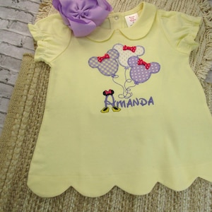 May include: A pale yellow short-sleeved shirt with a scalloped hem and a Peter Pan collar. The front features an embroidered design of three mouse-ear balloons with red bows and the name Amanda in purple text with mouse shoes below it.