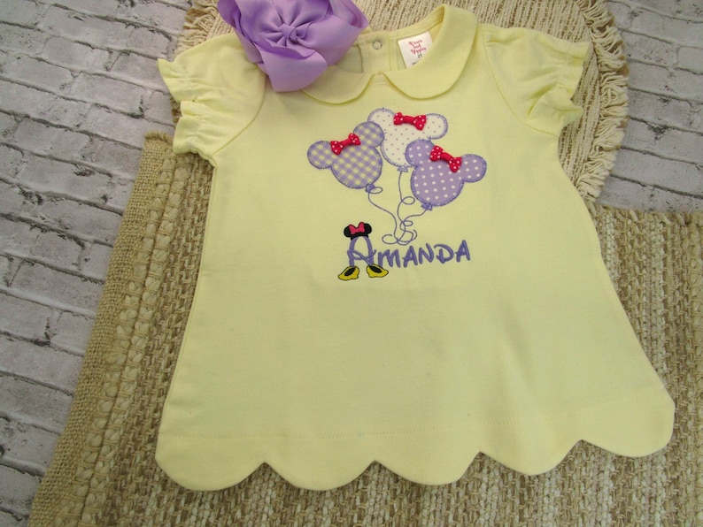 May include: A pale yellow short-sleeved shirt with a scalloped hem and a Peter Pan collar. The front features an embroidered design of three mouse-ear balloons with red bows and the name Amanda in purple text with mouse shoes below it.