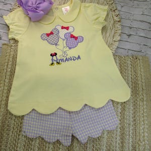 May include: A pale yellow short-sleeved shirt with a scalloped hem and a Peter Pan collar, featuring an embroidered design of three mouse-shaped balloons with red bows and the name Amanda in purple text, paired with light purple gingham shorts.