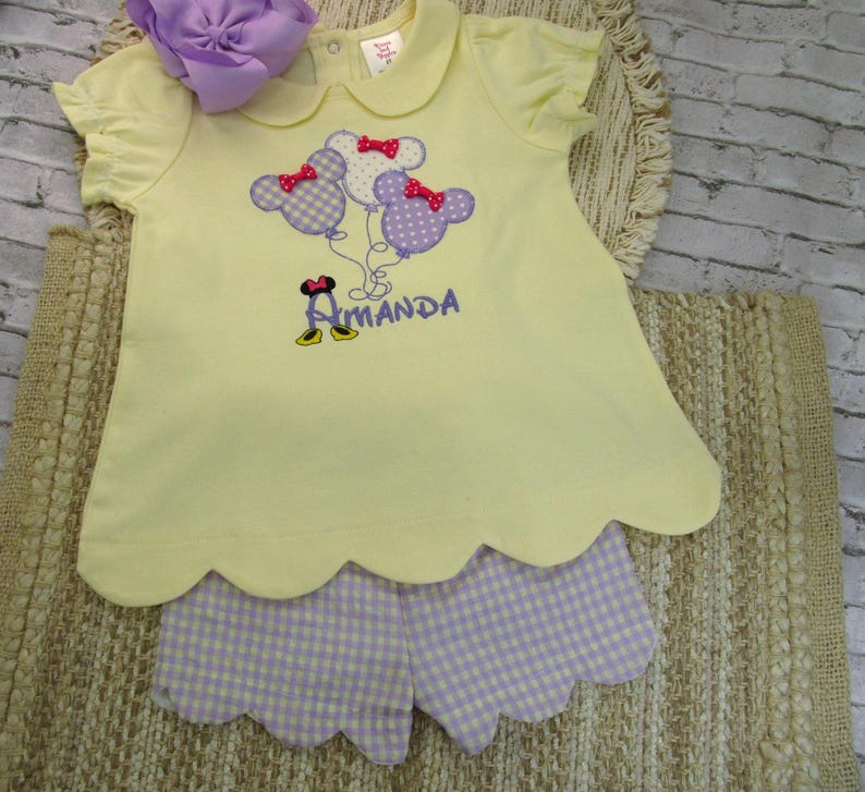 May include: A pale yellow short-sleeved shirt with a scalloped hem and a Peter Pan collar, featuring an embroidered design of three mouse-shaped balloons with red bows and the name Amanda in purple text, paired with light purple gingham shorts.