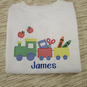 May include: White t-shirt with an embroidered design of a train carrying school supplies, including a book, scissors, crayons, and two red apples. The name "James" is embroidered below the train.