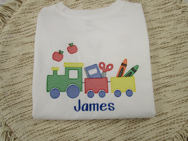 May include: White t-shirt with an embroidered design of a train carrying school supplies, including a book, scissors, crayons, and two red apples. The name "James" is embroidered below the train.