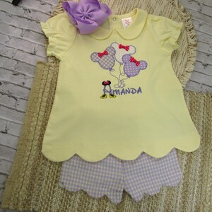 May include: A pale yellow short-sleeved shirt with a scalloped hem and a purple gingham pair of shorts. The shirt features an embroidered design of three mouse-shaped balloons with red bows and the name Amanda in purple thread with yellow shoes.