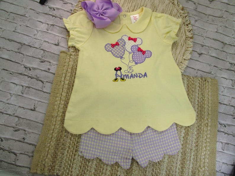 May include: A pale yellow short-sleeved shirt with a scalloped hem and a purple gingham pair of shorts. The shirt features an embroidered design of three mouse-shaped balloons with red bows and the name Amanda in purple thread with yellow shoes.