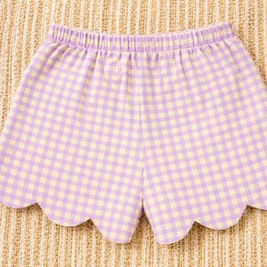 May include: Shorts featuring a light purple and pale yellow gingham check pattern. The garment has an elasticized waistband and a scalloped hemline along the bottom edge, creating a curved, decorative finish on each leg opening.