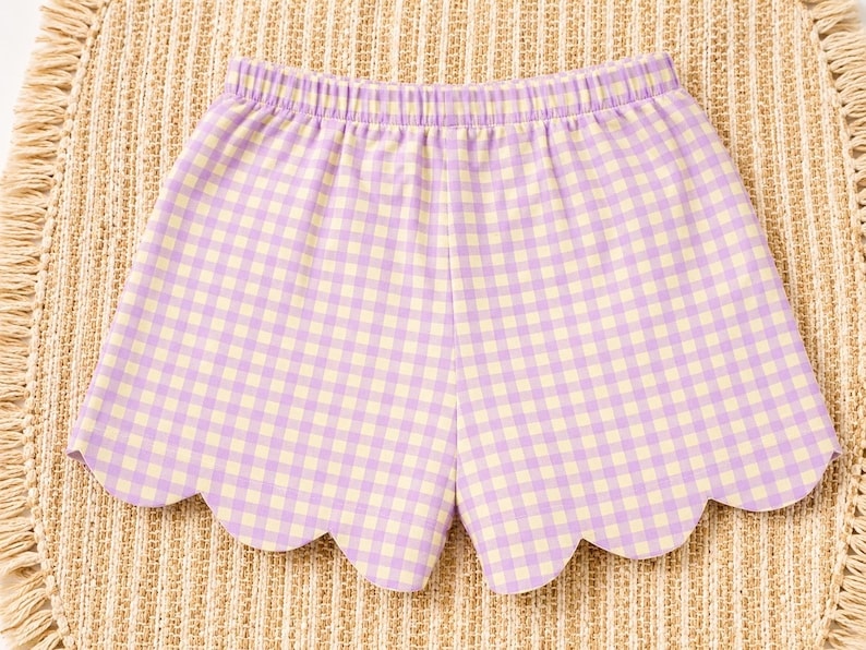 May include: Shorts featuring a light purple and pale yellow gingham check pattern. The garment has an elasticized waistband and a scalloped hemline along the bottom edge, creating a curved, decorative finish on each leg opening.