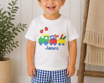 May include: A white short-sleeved t-shirt featuring a colorful embroidered train graphic carrying school supplies and two red apples, with the name James embroidered in blue below. The shirt is paired with blue and white gingham shorts.