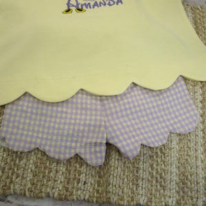 May include: A pale yellow top with a scalloped hem and the embroidered name Amanda in purple, paired with light purple and white gingham shorts featuring a matching scalloped hemline. The set displays a clean, simple design with embroidery.