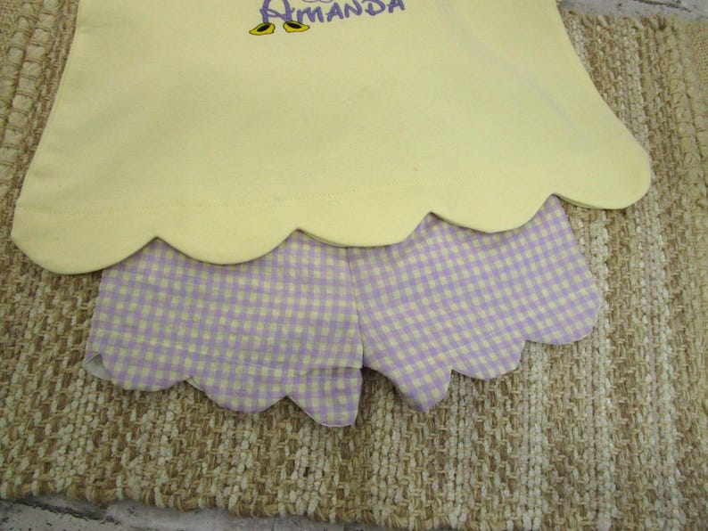 May include: A pale yellow top with a scalloped hem and the embroidered name Amanda in purple, paired with light purple and white gingham shorts featuring a matching scalloped hemline. The set displays a clean, simple design with embroidery.