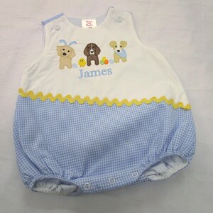 May include: A white and blue gingham baby romper with embroidered dog and chick designs. The romper has a yellow scalloped trim and the name "James" embroidered in blue.