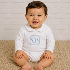 May include: A white long-sleeved baby bodysuit featuring a rounded Peter Pan collar and a light blue embroidered square frame on the chest containing the monogram letters SBA in a classic serif font, centered on the front of the garment.