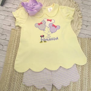 May include: A pale yellow short-sleeved shirt with a Peter Pan collar and scalloped hem, featuring an embroidered design of three polka-dot mouse-ear balloons and the name Amanda. Paired with light purple and white gingham scalloped shorts.