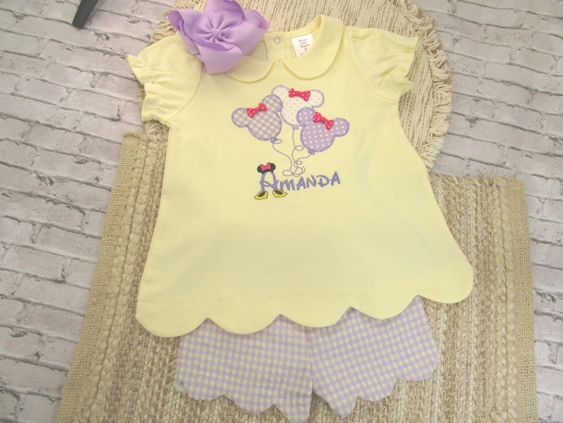 May include: A pale yellow short-sleeved shirt with a Peter Pan collar and scalloped hem, featuring an embroidered design of three polka-dot mouse-ear balloons and the name Amanda. Paired with light purple and white gingham scalloped shorts.
