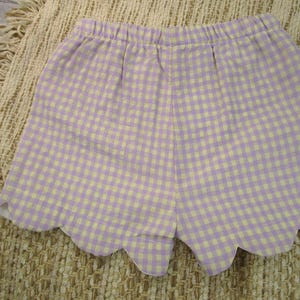 May include: A pair of shorts featuring a light purple and white gingham check pattern. The garment has an elasticized waistband and a scalloped hemline along the bottom edge, creating a distinct curved finish to the leg openings of the shorts.
