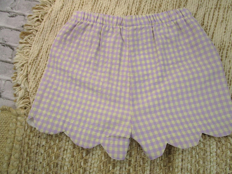 May include: A pair of shorts featuring a light purple and white gingham check pattern. The garment has an elasticized waistband and a scalloped hemline along the bottom edge, creating a distinct curved finish to the leg openings of the shorts.