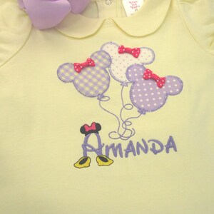 May include: A pale yellow short-sleeved shirt with a Peter Pan collar and ruffled sleeves. The front features an embroidered design of three mouse-shaped balloons in purple gingham and polka dots, each with a small red bow, and the name Amanda.