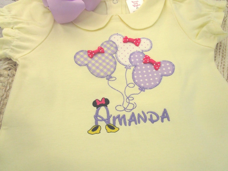 May include: A pale yellow short-sleeved shirt with a Peter Pan collar and ruffled sleeves. The front features an embroidered design of three mouse-shaped balloons in purple gingham and polka dots, each with a small red bow, and the name Amanda.