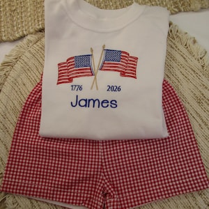 May include: A white t-shirt and red gingham shorts set. The shirt has embroidered American flags, the year 1776, and the name "James" in blue. The shorts are a red and white checkered pattern, suitable for patriotic events.