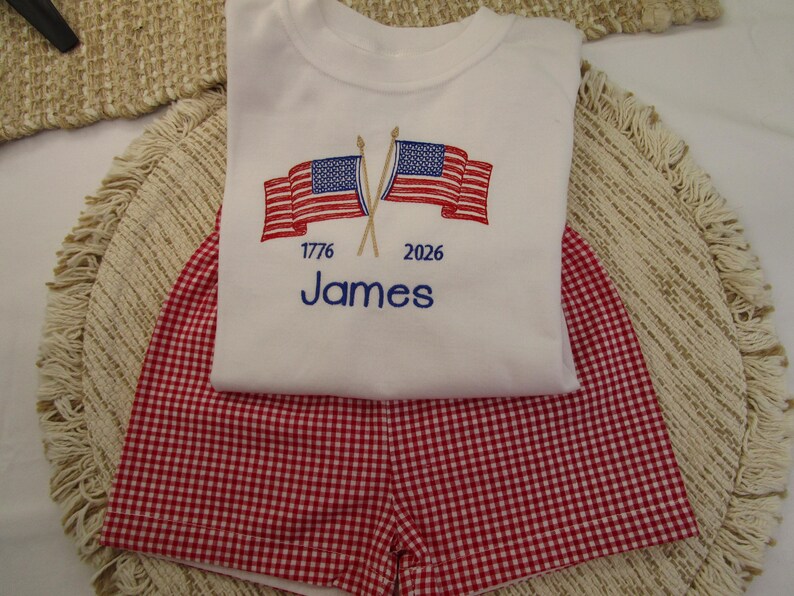 May include: A white t-shirt and red gingham shorts set. The shirt has embroidered American flags, the year 1776, and the name "James" in blue. The shorts are a red and white checkered pattern, suitable for patriotic events.