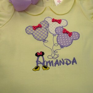 May include: A pale yellow short-sleeved shirt with a Peter Pan collar. The front features an embroidered design of three Mickey Mouse-shaped balloons in purple gingham and polka dot patterns, each with a red bow, above the name Amanda in purple.