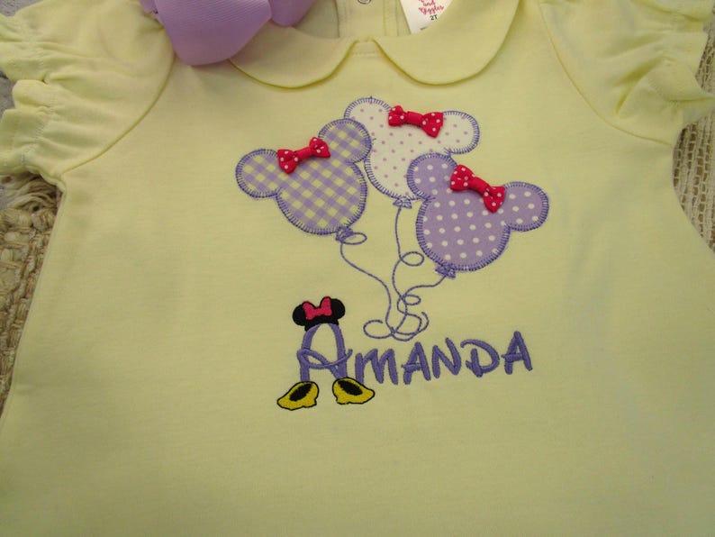 May include: A pale yellow short-sleeved shirt with a Peter Pan collar. The front features an embroidered design of three Mickey Mouse-shaped balloons in purple gingham and polka dot patterns, each with a red bow, above the name Amanda in purple.