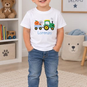 May include: A white short-sleeved t-shirt featuring a graphic of a green tractor pulling a blue trailer filled with a smiling orange pumpkin and a white ghost. The name Lorenzo is printed in light blue text below the tractor graphic.