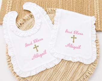 May include: A white baby bib and a white burp cloth, both featuring ruffled edges. Each item is embroidered with the pink text God Bless and Abigail, separated by a gold-colored cross symbol centered between the two lines of text.