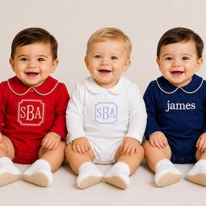 May include: Three long-sleeve baby bodysuits with Peter Pan collars and white trim. The red and white bodysuits feature a centered monogram with the letters SBA in a decorative frame. The navy blue bodysuit features the name james in white text.