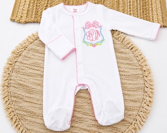 May include: A white long-sleeved footed sleeper with pink trim along the neckline, cuffs, and front button placket. The chest features an embroidered crest with a pink plaid bow, pink monogrammed letters, and a small floral design at the base.