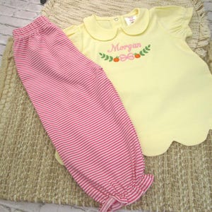 May include: A pale yellow short-sleeved shirt with a rounded collar and a scalloped hem, featuring an embroidered design of green leaves, orange pumpkins, a pink bow, and the name Morgan in pink script, paired with pink and white striped pants.