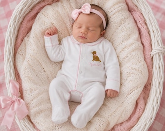 Puede incluir: Un pijama blanco de manga larga con pies, ribetes rosas y una diadema rosa con un lazo. El pijama tiene un gráfico bordado de un perro marrón y el nombre Karter cosido con hilo rosa en el pecho, usado por un bebé durmiendo.