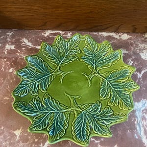May include: A green ceramic plate with a leaf design. The plate is round with a scalloped edge and features a raised relief pattern of leaves arranged in a circular pattern. The plate is a vibrant shade of green.