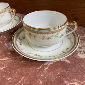 May include: A set of two white teacups and saucers with a delicate floral pattern and gold trim. The cups have a rounded shape and a small handle. The saucers have a scalloped edge.