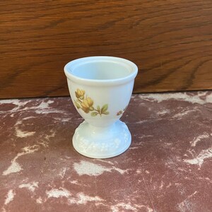 May include: White ceramic egg cup with a floral design featuring yellow flowers and green leaves. The cup has a pedestal base with a textured design.