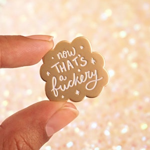 A brown hand holds up a matte gold enamel pin says, now thats a fuckery, in glittery white enamel with little white stars surrounding the letters.