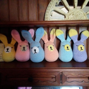 May include: Six crocheted bunny plushes in different colors, including yellow, pink, blue, and purple. The bunnies have black eyes and noses and are arranged in a row on a wooden shelf.