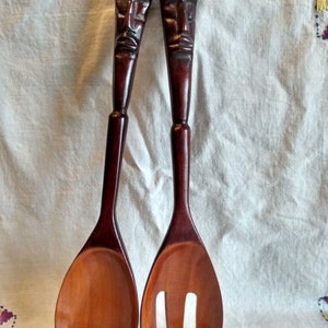 May include: A set of two wooden salad servers with carved wooden faces. The server on the left is a spoon and the server on the right is a fork.