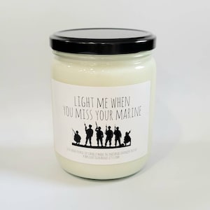 May include: A white candle in a glass jar with a black lid. The label reads "Light me when you miss your marine" and features a silhouette of a group of soldiers.
