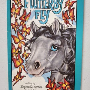May include: A book cover with a blue background and a gray horse with blue eyes. The title of the book is "Flutterby Fly" and the author is Stephen Cosgrove. The book is illustrated by Robin James. The cover is decorated with colorful butterflies.