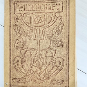 May include: A tan-colored book cover featuring the word WILDCRAFT embossed in a rectangular frame at the top. Below the text is a detailed brown line-art illustration of a large central flower with leaves and intricate, swirling vine patterns.