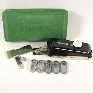 以下が含まれることがあります： Singerのミシンアタッチメントキット。黒とシルバーのミシンアタッチメント、緑色のケース、およびいくつかのシルバーとグレーのアタッチメントが含まれています。Singerのロゴは、ミシンアタッチメントとケースに表示されています。